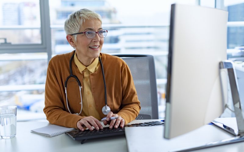 Photo of happy female Senior doctor working at desk with a computer in the office during the day. Smiling Beautiful friendly senior grey-haired lady posing in an office looking at PC computer with a lovely smile.