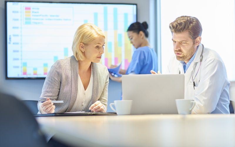 male doctor and female businesswoman chat over a laptop as a young nurse collates info on a smart board